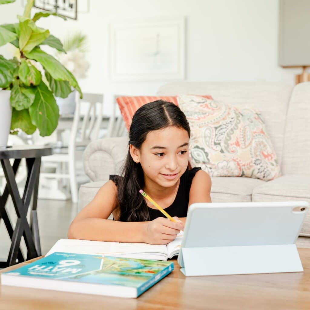 Photograph of Girl Watching Homeschool Math Lesson on a Tablet Whilst Writing in her Math Course Book for Grade 6